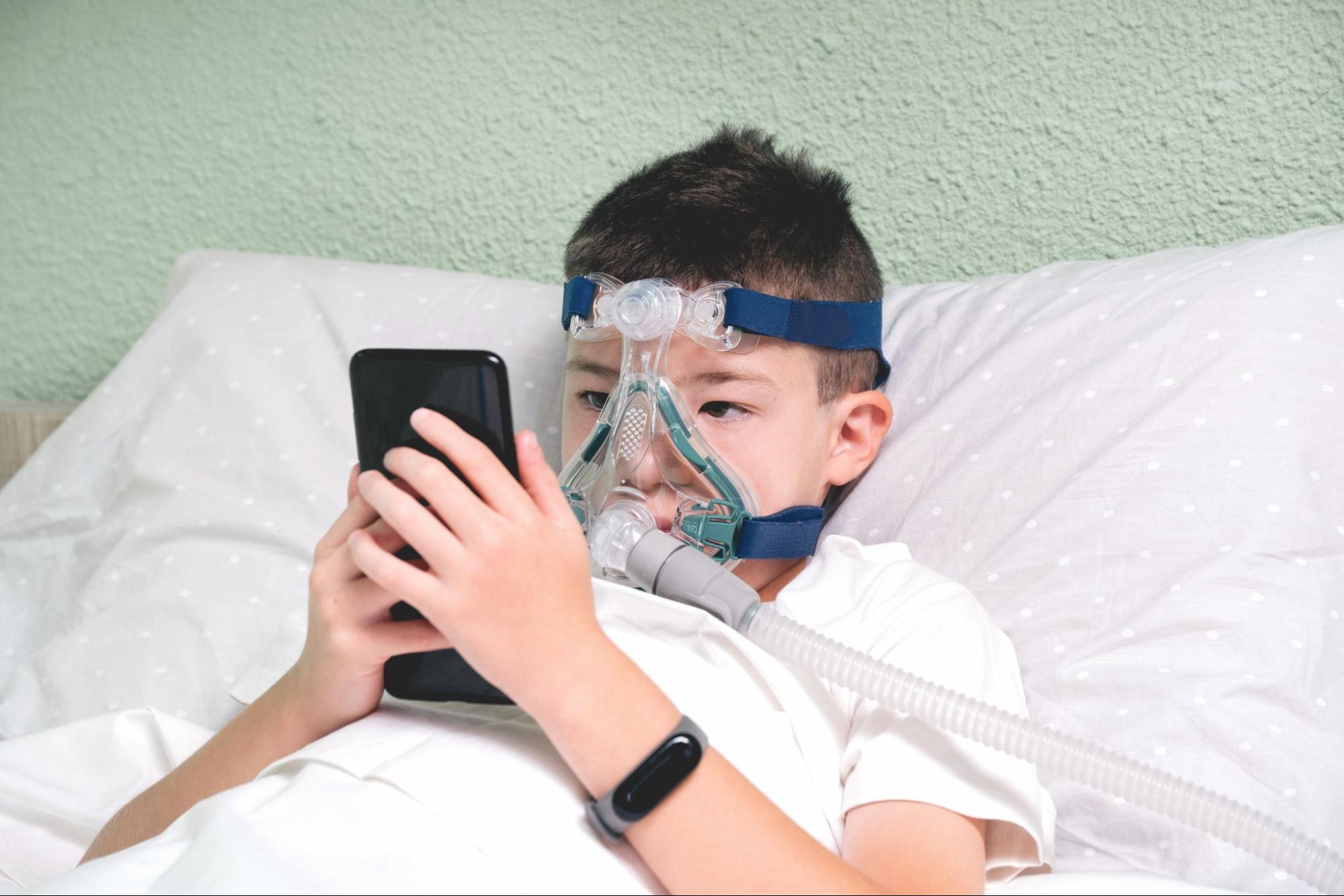 A child resting in bed while comfortably wearing a full face CPAP mask and using a mobile device.