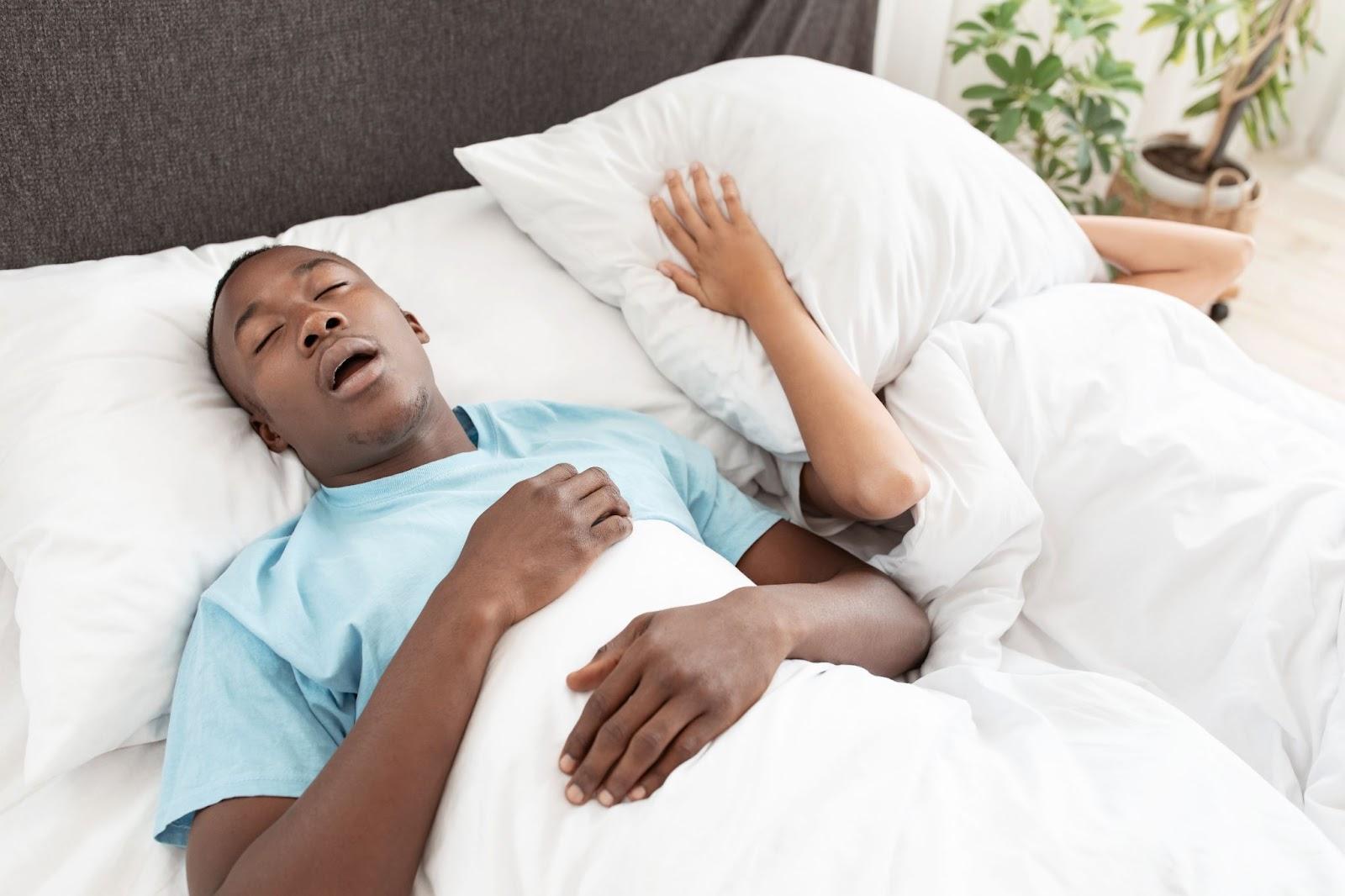 Man snoring loudly in bed while partner covers ears with a pillow, showing common sleep apnea symptoms.
