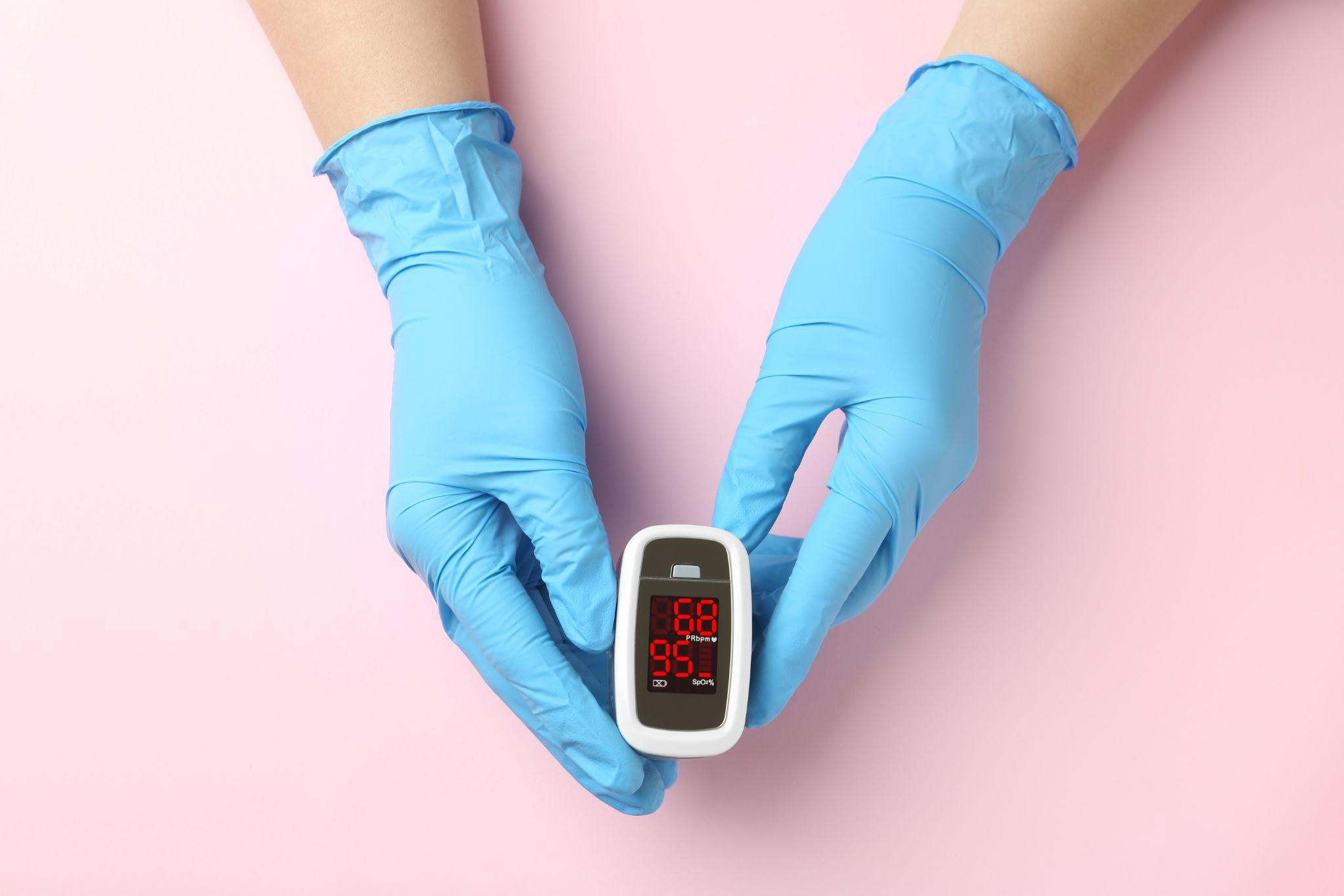 Hands in medical gloves holding a fingertip pulse oximeter, a respiratory monitoring device offered by a medical supply company.