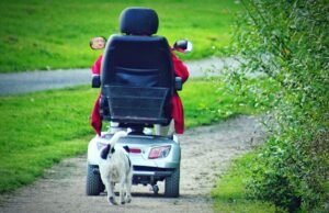 Senior riding an electric scooter for elderly mobility on a park path while a dog walks beside the scooter.