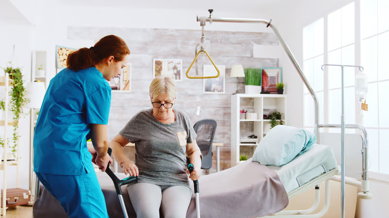 Caregiver assisting an older adult beside a hospital bed with patient lift equipment available for safer transfers.