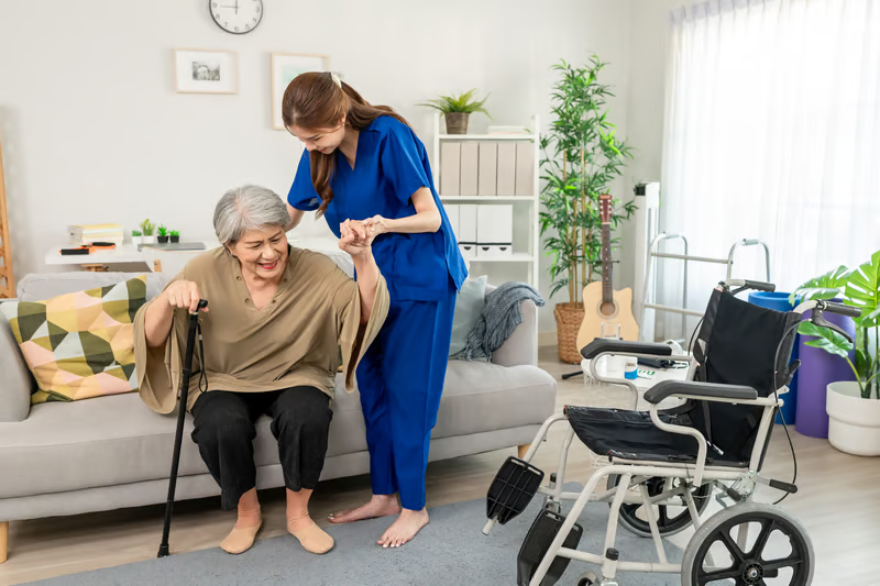 Caregiver helping an older adult stand at home near a wheelchair and walker, showing how home care equipment can support safer daily mobility.